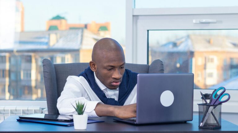 Adult male focused on work at his desk with a laptop in a modern office setting.